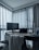 A modern home office setup with a sleek desk positioned by large windows with sheer curtains. The desktop surface includes a monitor, a closed laptop, a desk lamp, and a small figurine. A chair with a blanket draped over the back is in front of the desk, and the overall setting appears organized and minimalist.