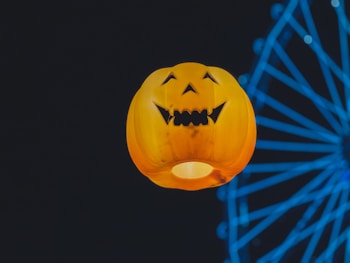 A glowing jack-o'-lantern with a classic carved face is set against a dark background with the structure of a Ferris wheel illuminated in blue light visible in the background.