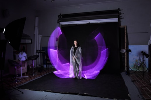 A woman is standing in a photography studio, wearing a long striped dress. There is a dynamic, purple light painting effect that forms a semi-circular pattern around her. The studio setup includes lighting equipment and a dark backdrop. To the side, there are stools and a fan as part of the studio arrangement.