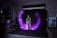 A woman is standing in a photography studio, wearing a long striped dress. There is a dynamic, purple light painting effect that forms a semi-circular pattern around her. The studio setup includes lighting equipment and a dark backdrop. To the side, there are stools and a fan as part of the studio arrangement.