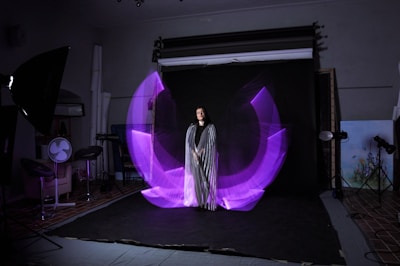 A woman is standing in a photography studio, wearing a long striped dress. There is a dynamic, purple light painting effect that forms a semi-circular pattern around her. The studio setup includes lighting equipment and a dark backdrop. To the side, there are stools and a fan as part of the studio arrangement.