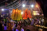 A vibrant and crowded outdoor festival scene with large, colorful decorative structures illuminated by bright lights. Numerous people gather around, many wearing traditional attire, creating a lively and festive atmosphere. Overhead decorations and bright lighting add to the celebratory ambiance.