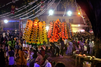 A vibrant and crowded outdoor festival scene with large, colorful decorative structures illuminated by bright lights. Numerous people gather around, many wearing traditional attire, creating a lively and festive atmosphere. Overhead decorations and bright lighting add to the celebratory ambiance.