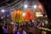 A vibrant and crowded outdoor festival scene with large, colorful decorative structures illuminated by bright lights. Numerous people gather around, many wearing traditional attire, creating a lively and festive atmosphere. Overhead decorations and bright lighting add to the celebratory ambiance.