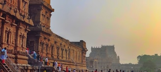 A group of people is gathered near an ancient stone temple with intricate carvings and architecture. The temple structure is detailed with pillars and ornate designs, characteristic of historical Indian architecture. The ambient lighting suggests a setting sun, casting a warm glow over the scene.