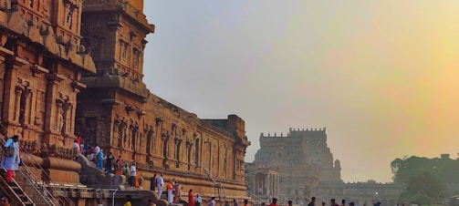 A group of people is gathered near an ancient stone temple with intricate carvings and architecture. The temple structure is detailed with pillars and ornate designs, characteristic of historical Indian architecture. The ambient lighting suggests a setting sun, casting a warm glow over the scene.