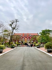 A large, historic red building with white accents serves as the backdrop. In front of the building stands a prominent golden statue. The area is landscaped with paths and potted plants, and a group of people are gathered near the statue. Flags are positioned prominently atop the building.
