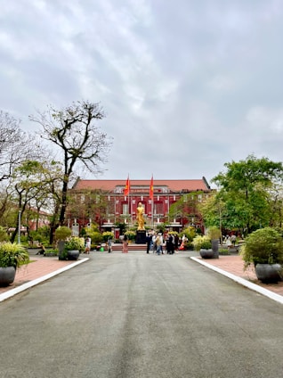 A large, historic red building with white accents serves as the backdrop. In front of the building stands a prominent golden statue. The area is landscaped with paths and potted plants, and a group of people are gathered near the statue. Flags are positioned prominently atop the building.