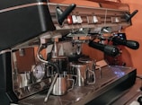 A professional espresso machine equipped with multiple handles and a pressure gauge. Several stainless steel milk frothing pitchers are arranged on the drip tray, while the machine's metallic components reflect the surrounding light.