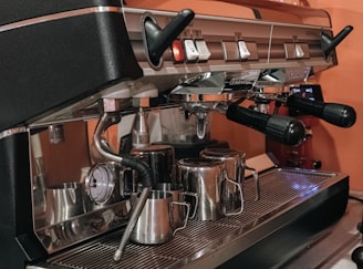 A professional espresso machine equipped with multiple handles and a pressure gauge. Several stainless steel milk frothing pitchers are arranged on the drip tray, while the machine's metallic components reflect the surrounding light.