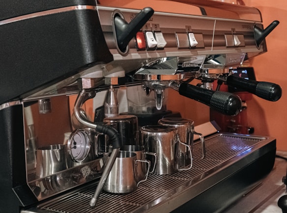 A professional espresso machine equipped with multiple handles and a pressure gauge. Several stainless steel milk frothing pitchers are arranged on the drip tray, while the machine's metallic components reflect the surrounding light.