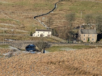 A rural landscape featuring two stone houses amidst a vast area of open grassland with sparse trees. A stone bridge crosses a small stream, with patches of snow on the ground. There are dry stone walls and rolling hills in the background.