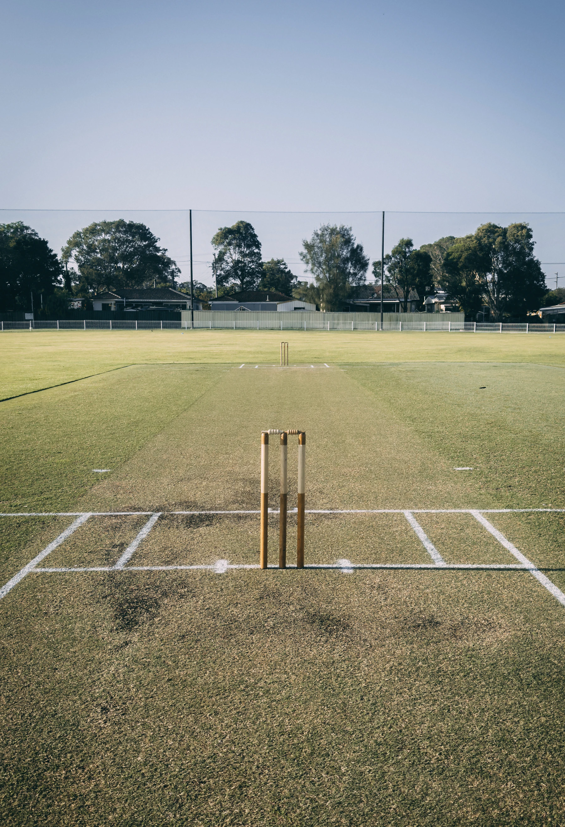 A mown cricket pitch with stumps at the near end, trees and a pavilion beyond