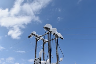 Snow-covered electrical power lines and poles against a clear blue sky with scattered white clouds.