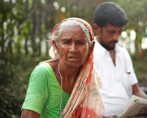 An elderly woman with grey hair and a weathered expression sits outdoors wearing a green dress and a patterned scarf on her head. Behind her, a younger man wearing a white shirt is sitting, holding a newspaper. The background is blurred with greenery, suggesting a park or natural setting.
