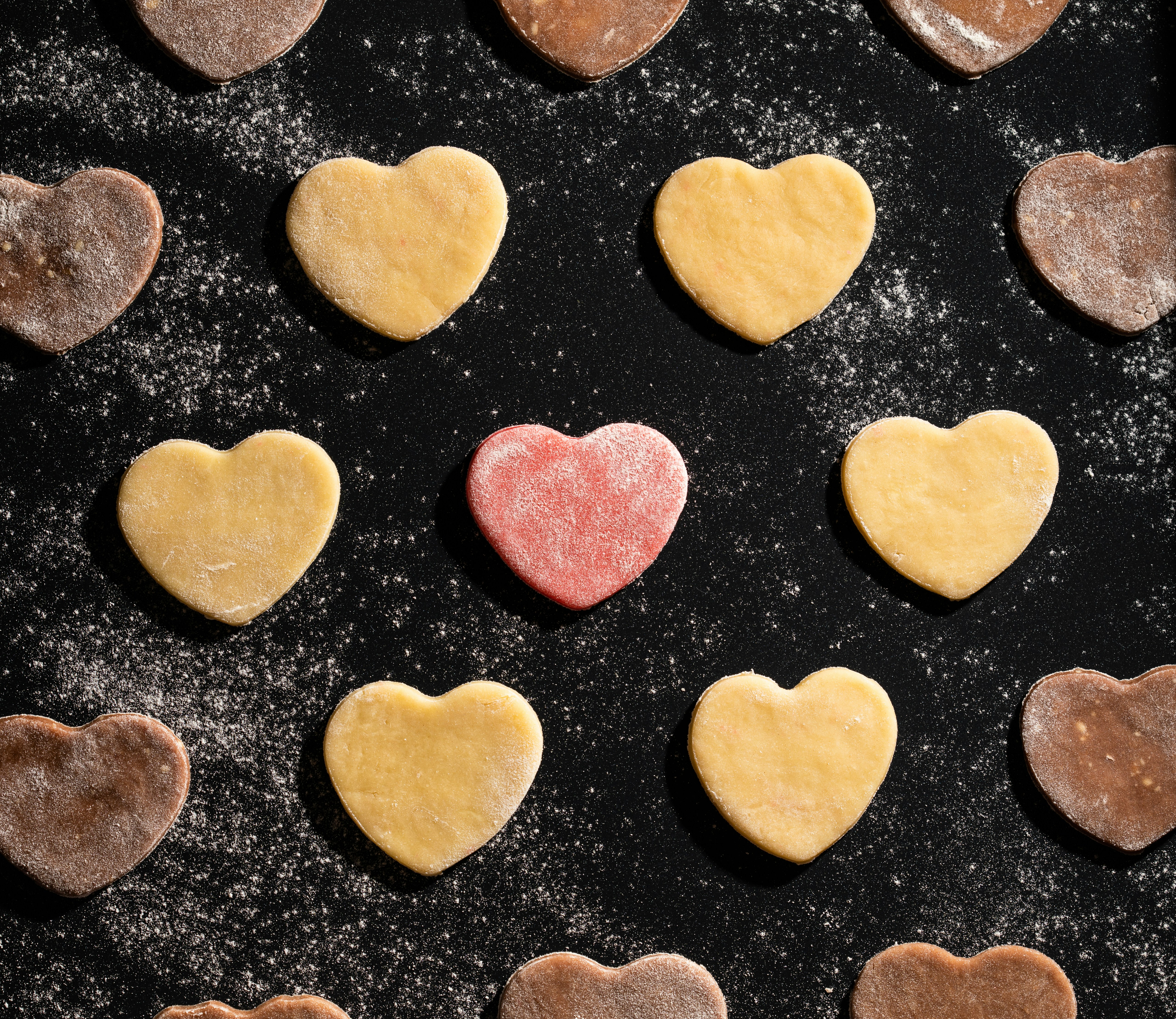 Macro view of pre-baked vanilla, pink and chocolate cookies in shape of hearts dusted with flour on black baking sheet