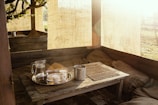 A rustic outdoor seating area bathed in warm sunlight, featuring a wooden table with a metal tray holding a glass pitcher and decorative paper cups. Burlap sacks and fabric panels hang around, creating a cozy, inviting atmosphere.