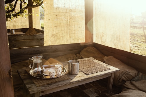 A rustic outdoor seating area bathed in warm sunlight, featuring a wooden table with a metal tray holding a glass pitcher and decorative paper cups. Burlap sacks and fabric panels hang around, creating a cozy, inviting atmosphere.
