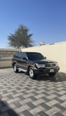 A dark-colored SUV is parked on a paved area under a clear blue sky. In the background, there is a beige wall with a sign in Arabic and English, and a tree is visible over the wall.