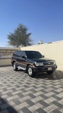 A dark-colored SUV is parked on a paved area under a clear blue sky. In the background, there is a beige wall with a sign in Arabic and English, and a tree is visible over the wall.