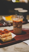 A wooden tray holds a dessert with layers of cream and cocoa powder next to a plastic cup filled with iced coffee, featuring a mix of milk and espresso. In the background, two ceramic mugs and a blurred orange-colored dessert can be seen.