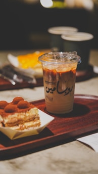 A wooden tray holds a dessert with layers of cream and cocoa powder next to a plastic cup filled with iced coffee, featuring a mix of milk and espresso. In the background, two ceramic mugs and a blurred orange-colored dessert can be seen.