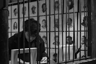A person is seated behind bars with a book in front of them. The background is filled with various black-and-white portraits pinned to a wall, depicting different people.