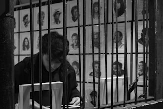 A person is seated behind bars with a book in front of them. The background is filled with various black-and-white portraits pinned to a wall, depicting different people.