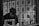 A person is seated behind bars with a book in front of them. The background is filled with various black-and-white portraits pinned to a wall, depicting different people.