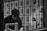 A person is seated behind bars with a book in front of them. The background is filled with various black-and-white portraits pinned to a wall, depicting different people.