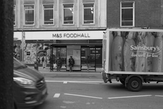 A busy urban street scene with a food hall on the left and a delivery truck on the right displaying a large advertisement. Several people are sitting at a bus stop in front of the food hall. The street is active with vehicles moving past.