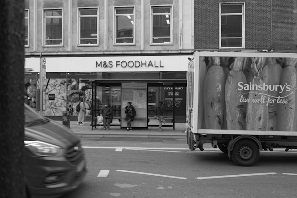A busy urban street scene with a food hall on the left and a delivery truck on the right displaying a large advertisement. Several people are sitting at a bus stop in front of the food hall. The street is active with vehicles moving past.