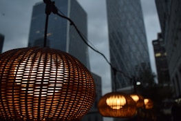 A series of warm, glowing woven lamps hanging in front of tall skyscrapers in an urban setting. The natural wicker texture of the lamps contrasts with the sleek glass and steel architecture in the background. The setting appears to be during twilight, with a moody, overcast sky.