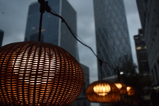 A series of warm, glowing woven lamps hanging in front of tall skyscrapers in an urban setting. The natural wicker texture of the lamps contrasts with the sleek glass and steel architecture in the background. The setting appears to be during twilight, with a moody, overcast sky.