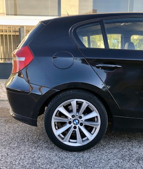 A close-up of the rear section of a black hatchback car, focusing on the rear wheel and tire. The car is parked on a paved street next to a building with metal fencing in the background. The wheel features a distinctive alloy rim with a recognizable logo at the center.