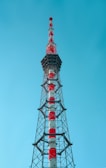 A tall, metal broadcast tower with sections painted in red and white stands against a clear blue sky. The structure is composed of a lattice framework and appears robust and industrial.