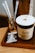A cup of coffee with a foamy top sits on a wooden tray next to a small glass with paper straws. The cup has a label that reads 'Daily Coffee Service'. Nearby, there is a packaged snack in shiny wrapping.