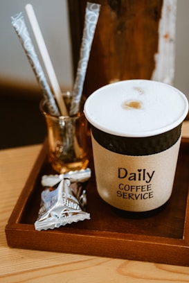 A cup of coffee with a foamy top sits on a wooden tray next to a small glass with paper straws. The cup has a label that reads 'Daily Coffee Service'. Nearby, there is a packaged snack in shiny wrapping.