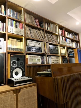 A well-organized wooden shelf filled with books, vinyl records, and vintage audio equipment, including speakers and a turntable. The shelf units have a warm, rustic appearance with a mix of modern and retro elements.