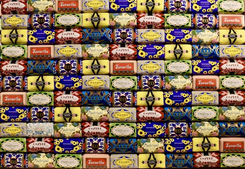 A neatly arranged collection of colorful soap bars, each with unique vintage packaging designs featuring flowers, geometric patterns, and various brand names. The soaps are stacked in rows, creating a visually appealing mosaic-like pattern with a retro aesthetic.
