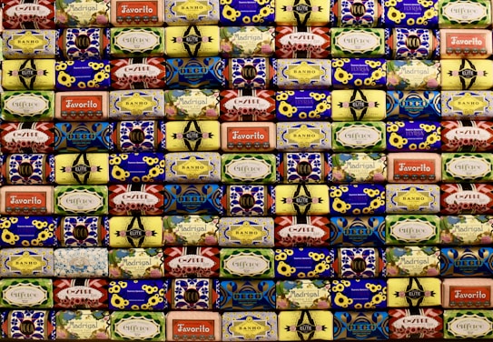 A neatly arranged collection of colorful soap bars, each with unique vintage packaging designs featuring flowers, geometric patterns, and various brand names. The soaps are stacked in rows, creating a visually appealing mosaic-like pattern with a retro aesthetic.