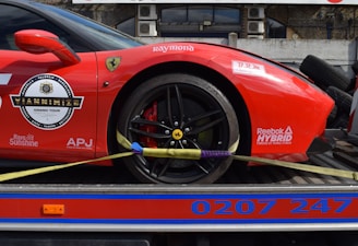 A red sports car with various sponsor logos and decals is securely fastened to a transport trailer. The focus is on the rear wheel and part of the body, showing the car's sleek design and high-performance tires.
