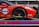 A red sports car with various sponsor logos and decals is securely fastened to a transport trailer. The focus is on the rear wheel and part of the body, showing the car's sleek design and high-performance tires.
