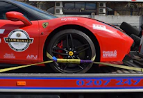 A red sports car with various sponsor logos and decals is securely fastened to a transport trailer. The focus is on the rear wheel and part of the body, showing the car's sleek design and high-performance tires.