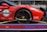 A red sports car with various sponsor logos and decals is securely fastened to a transport trailer. The focus is on the rear wheel and part of the body, showing the car's sleek design and high-performance tires.