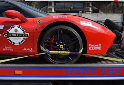 A red sports car with various sponsor logos and decals is securely fastened to a transport trailer. The focus is on the rear wheel and part of the body, showing the car's sleek design and high-performance tires.