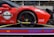 A red sports car with various sponsor logos and decals is securely fastened to a transport trailer. The focus is on the rear wheel and part of the body, showing the car's sleek design and high-performance tires.
