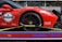 A red sports car with various sponsor logos and decals is securely fastened to a transport trailer. The focus is on the rear wheel and part of the body, showing the car's sleek design and high-performance tires.