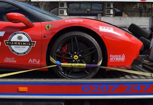 A red sports car with various sponsor logos and decals is securely fastened to a transport trailer. The focus is on the rear wheel and part of the body, showing the car's sleek design and high-performance tires.