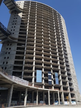 A large, unfinished high-rise building with a round structure is under construction. The building has multiple floors with concrete pillars and framework visible, indicating that it is in the early stages of development. Two sections of glass panels are installed on the lower levels. The upper floors are open and lack windows, giving a skeletal appearance. The sky is clear and blue, providing a backdrop to the construction site.